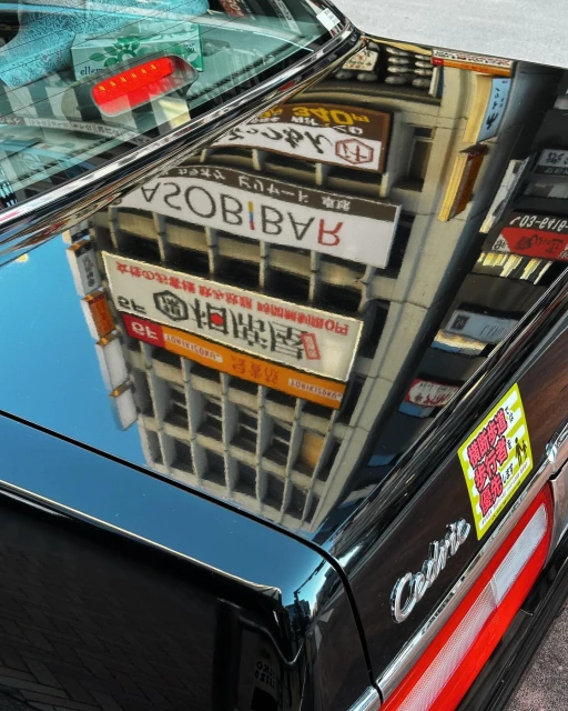 Reflection on a black car hood showing a building with signs in an urban environment, creating a surreal layered image.