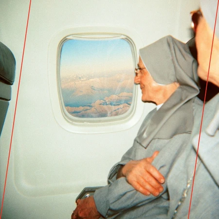 Nun in grey habit sits on airplane, looking out the window at clouds and mountains in the distance on a clear day, in-flight.