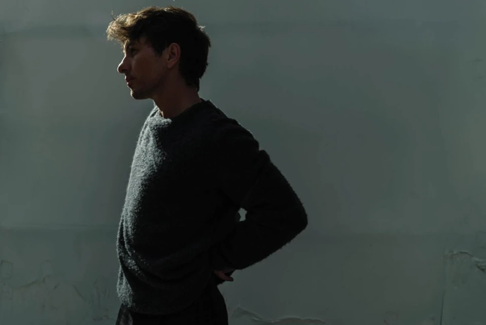 Profile of a young adult male in a dark sweater against a muted wall, bathed in directional light, casting strong shadows.