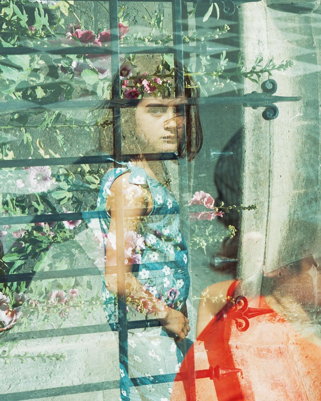 Young female model in a floral dress posing behind a metal gate, with a double exposure effect of flowers and foliage against a concrete wall.