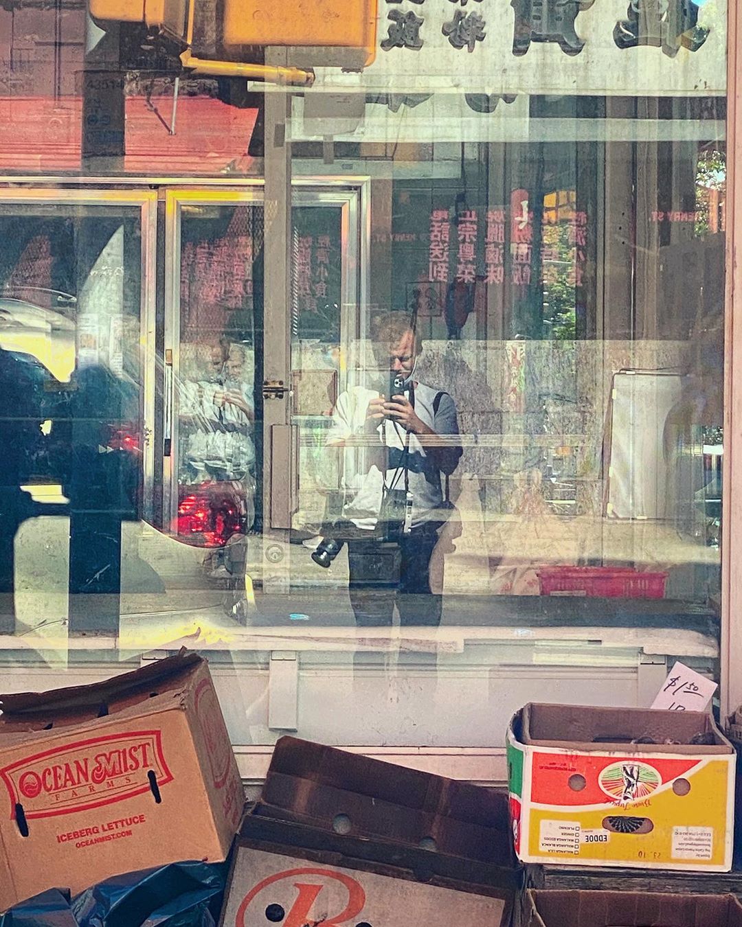 Reflection of a man photographing a storefront with produce boxes, including lettuce, stacked outside in a somewhat cluttered display.