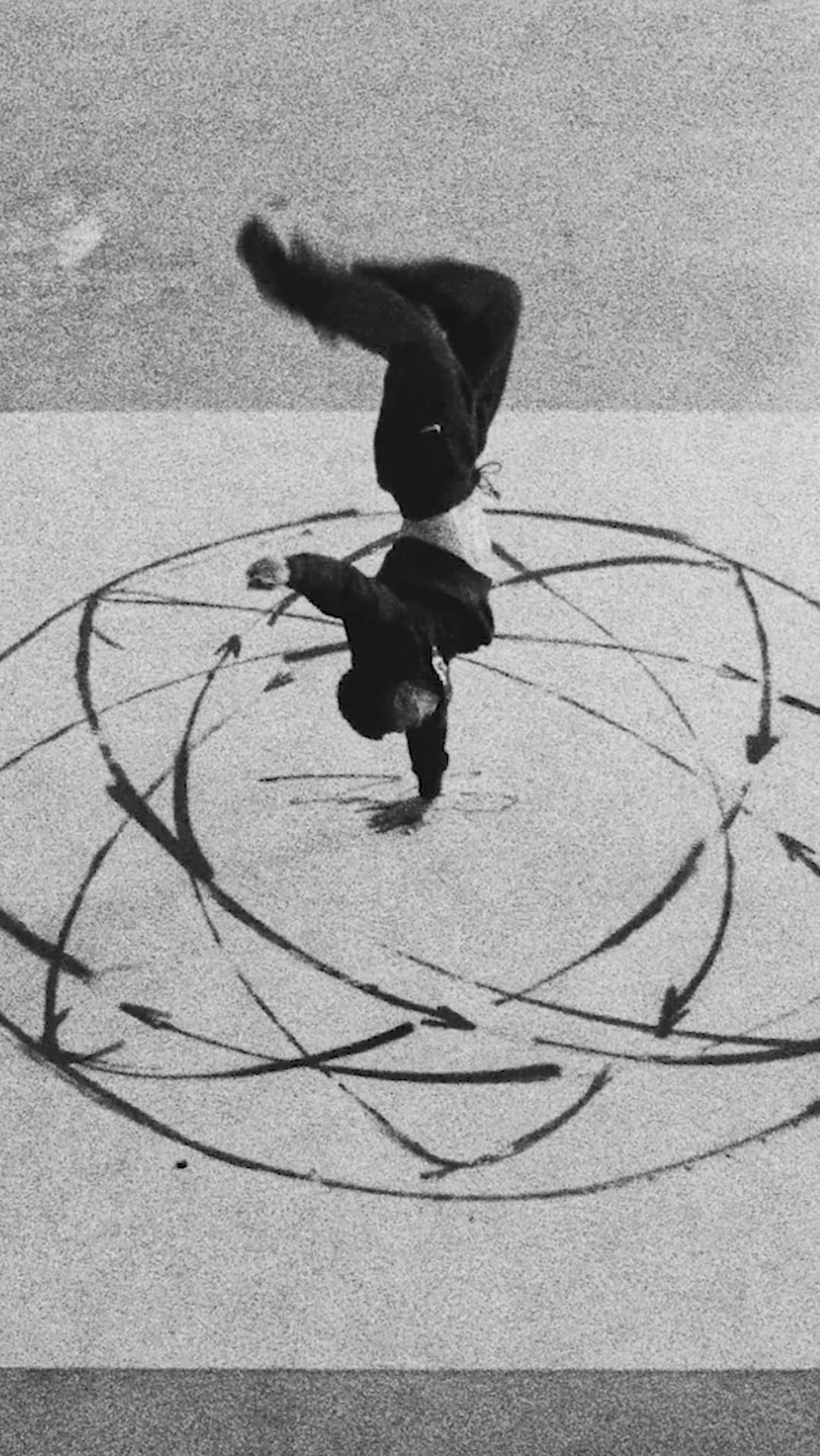A black and white photograph of a dancer balanced on one hand while upside down, performing on a surface with drawn circles.
