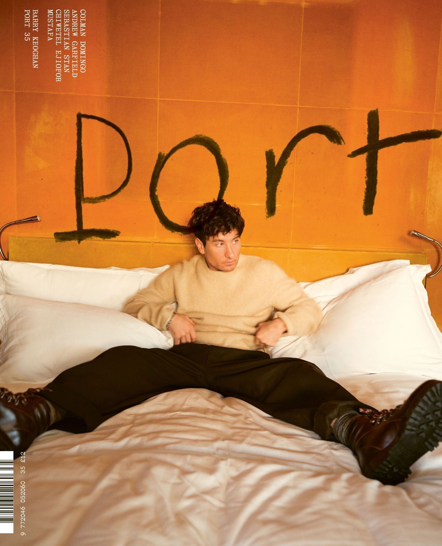 Man lounges on a bed with an orange wall featuring the word 'Port'. He is wearing a cream sweater, dark trousers, and brown boots.