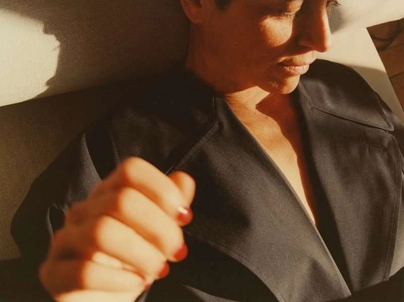 Close-up of a woman in a black trench coat with red nail polish lying in the sunlight, casting shadows on the surrounding surfaces.