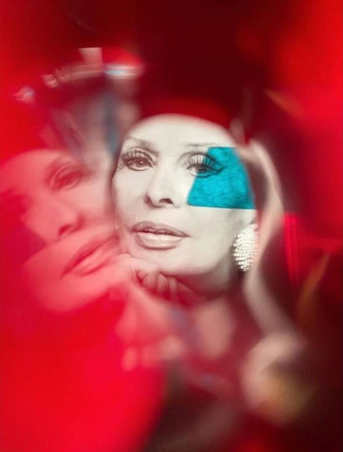 Portrait of a woman with blue art on her face and diamond earrings against a red background, with a blurred image of the same woman.