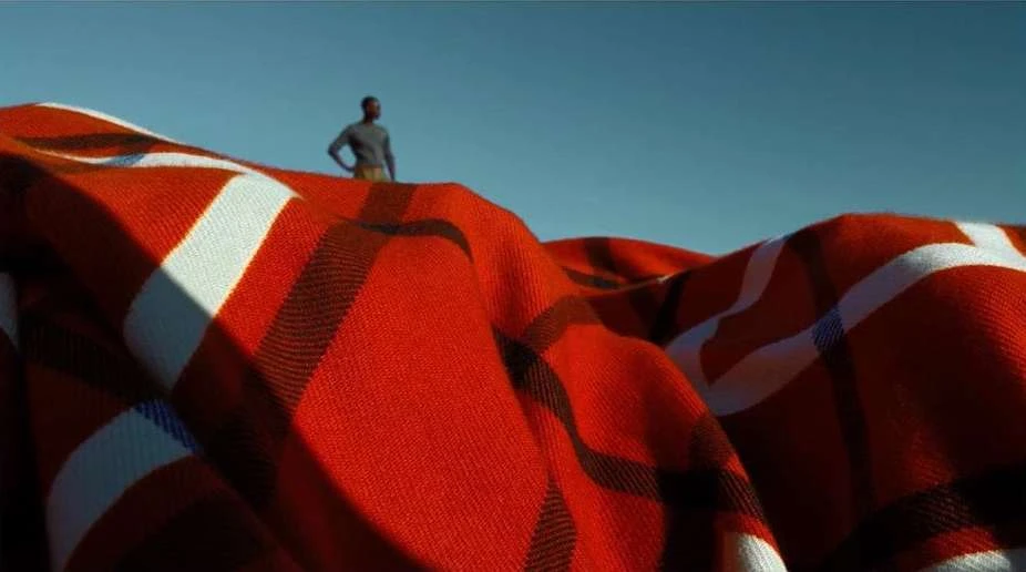 Man stands on a red patterned blanket in an outdoor setting against a clear blue sky, creating a surreal or miniature effect.