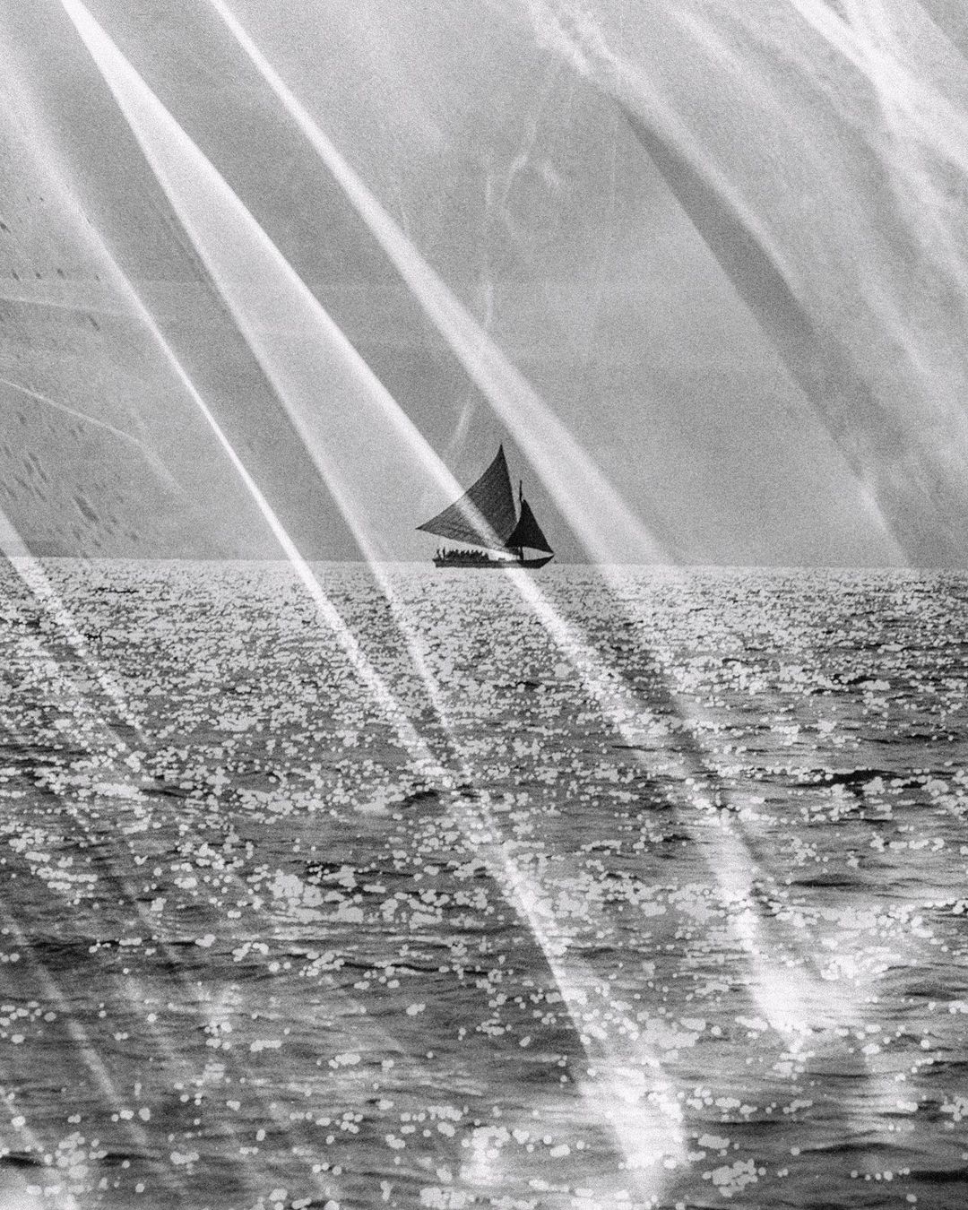 Black and white photograph of a sailboat on calm open water, light streaks across the frame, creating a vintage or artistic aesthetic.