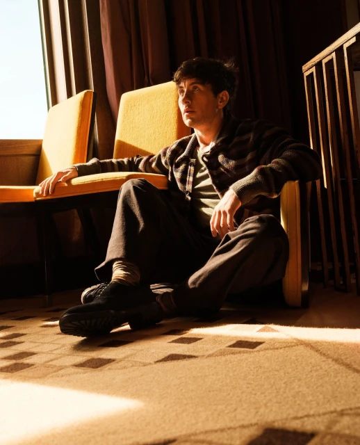 Adult male seated in mustard yellow chair with patterned rug. Sunlight and shadows create warm tones. Indoor residential scene.