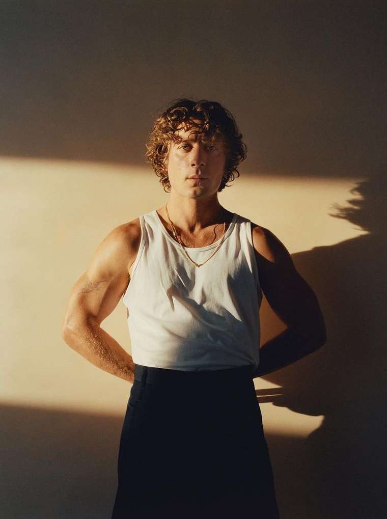 Portrait of a man with curly brown hair wearing a white tank top and dark trousers against a warm wall.