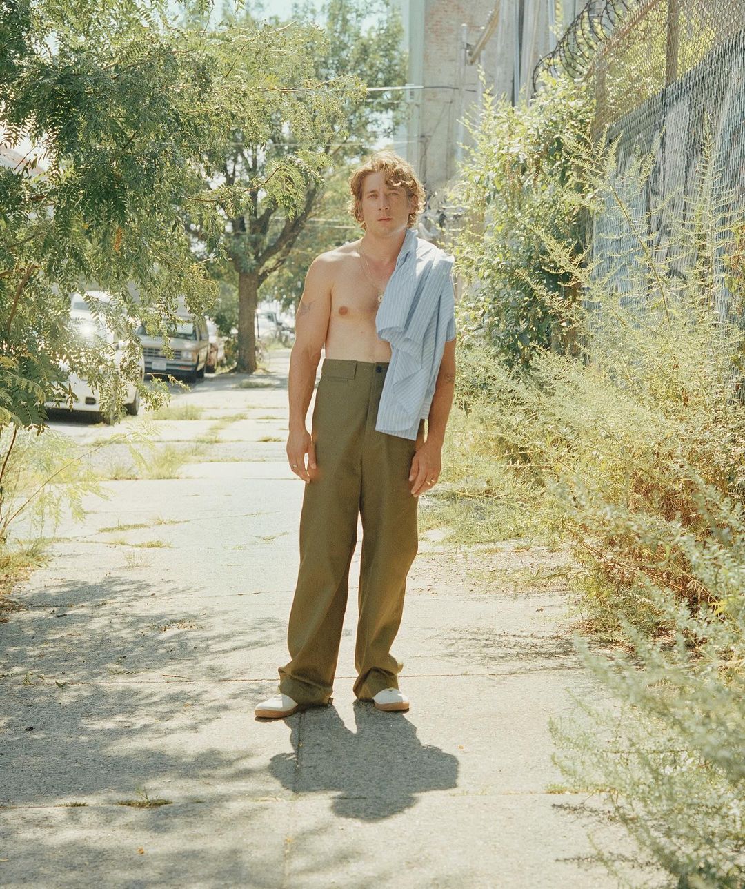 An adult male stands on a sidewalk in a suburban environment with a shirt slung over his shoulder, surrounded by greenery and buildings.