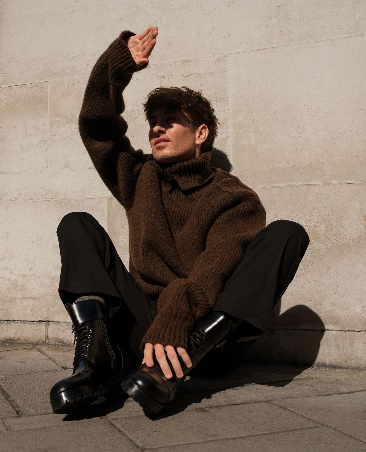 Young man in a brown sweater and black boots sits against a wall, holding his hand up to shield his face from the sunlight.