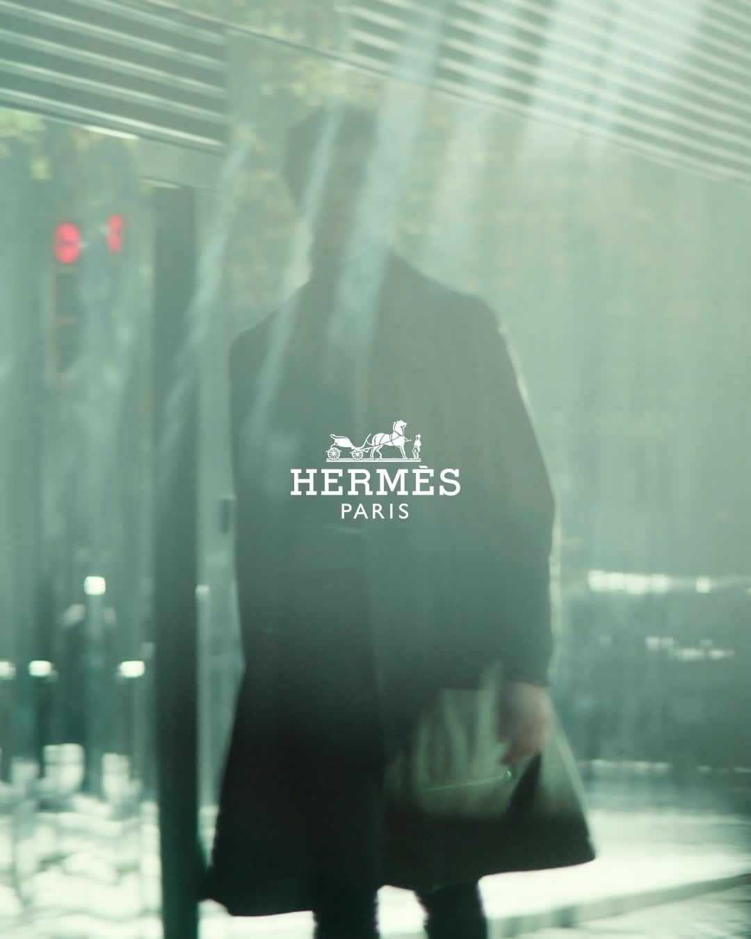 Fashion advertisement featuring a person walking with a handbag, showcasing the Hermes logo in an urban setting with soft lighting.