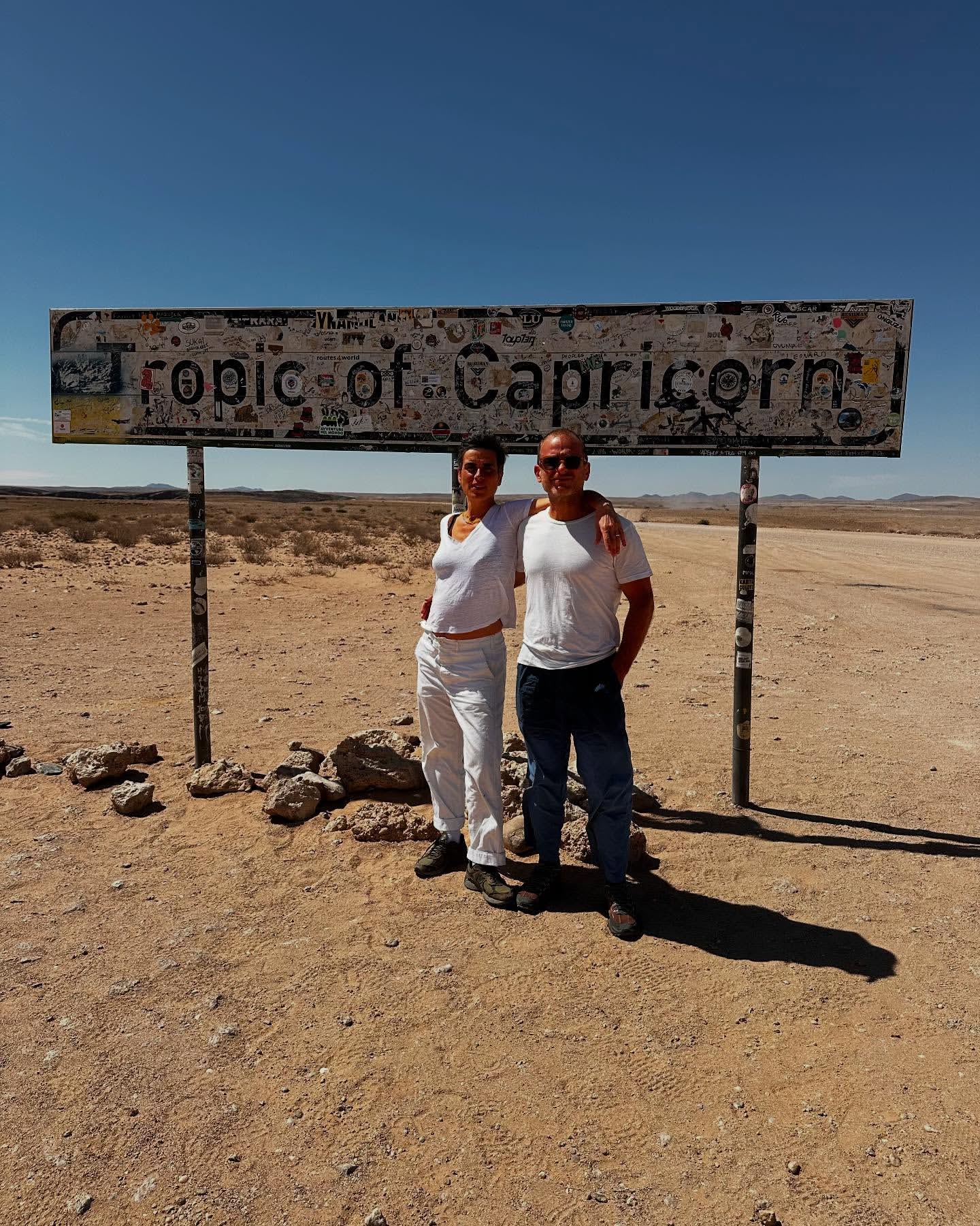 Couple poses at Tropic of Capricorn sign in desert landscape under clear sky, arid environment, horizon, and sunny day.