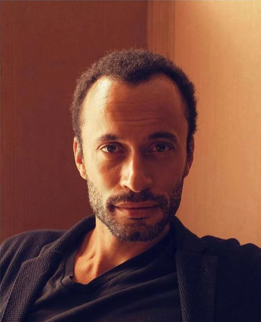 Portrait of a middle-aged adult male with short curly hair and a beard, wearing a black t-shirt and blazer, against a neutral backdrop.