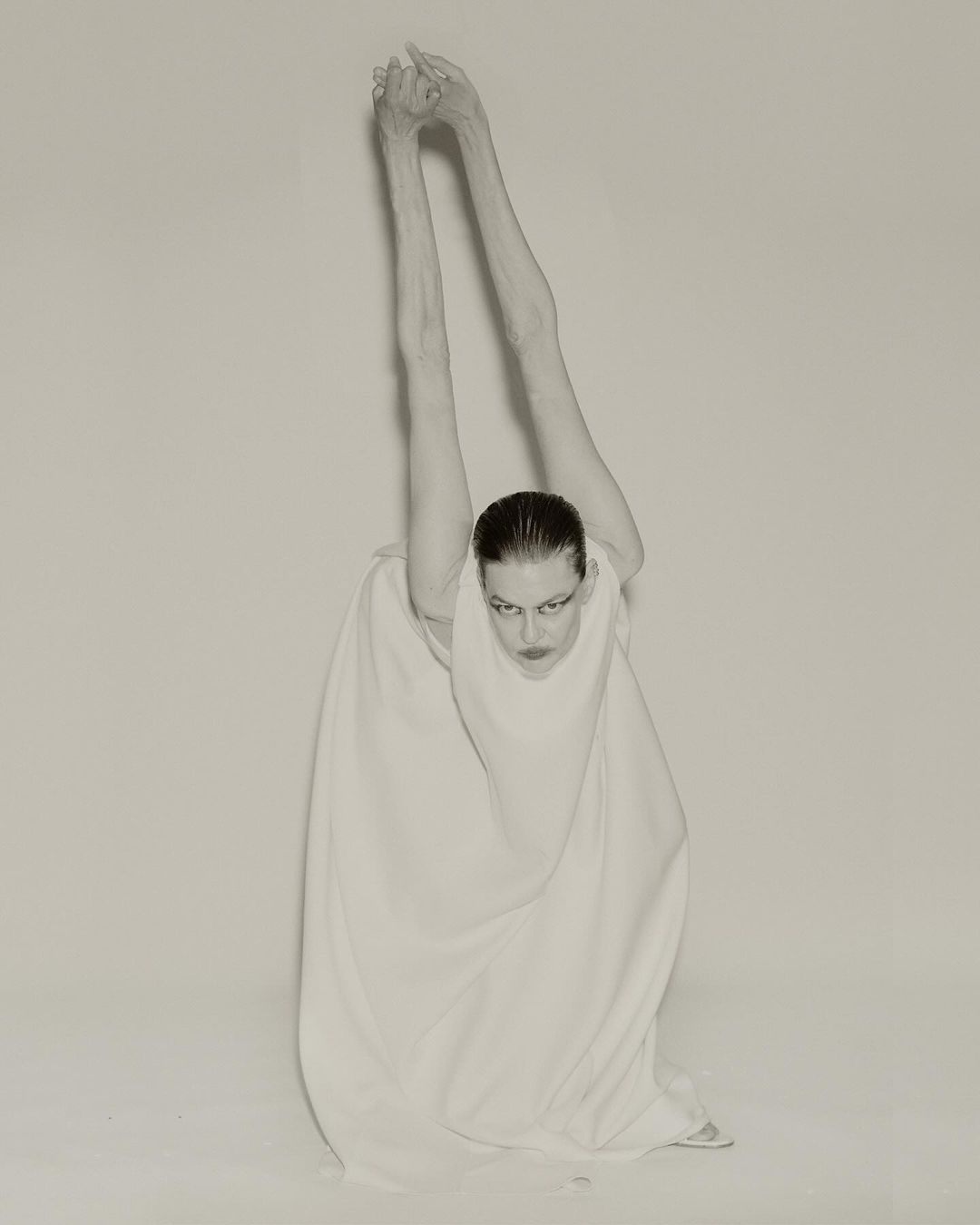 Monochrome studio portrait of a slender adult woman in a white dress with long arms raised overhead, fingers interlaced, gazing downwards.