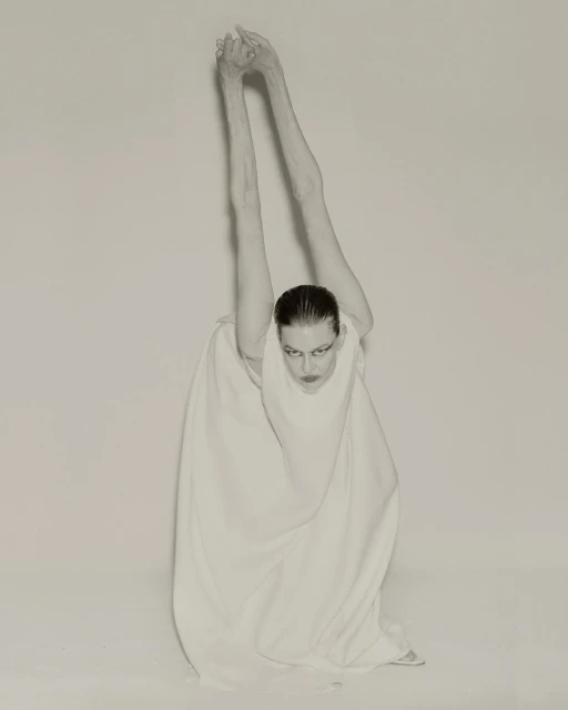 Monochrome studio portrait of a slender adult woman in a white dress with long arms raised overhead, fingers interlaced, gazing downwards.