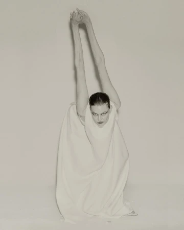 Monochrome studio portrait of a slender adult woman in a white dress with long arms raised overhead, fingers interlaced, gazing downwards.