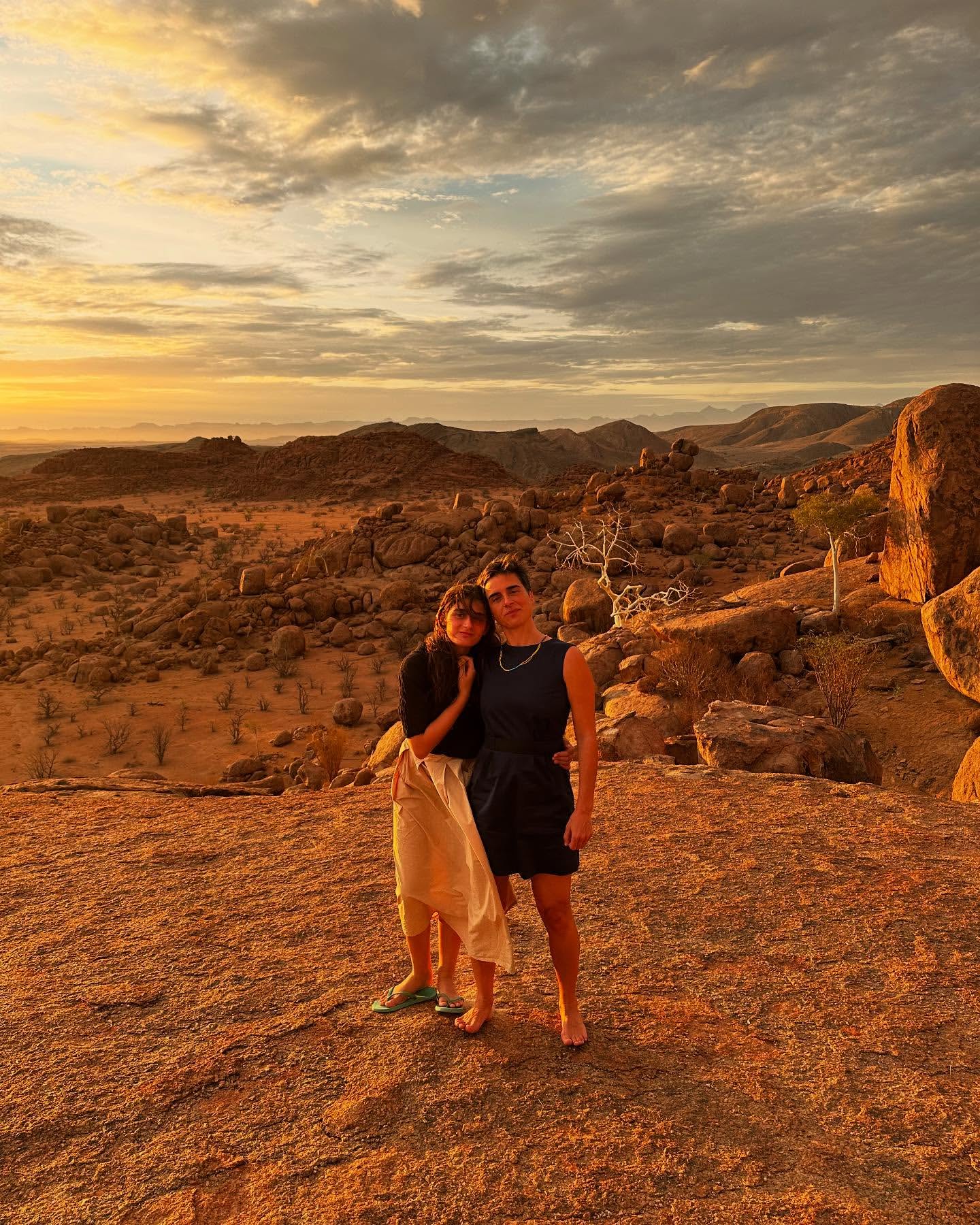 Two people pose on a rocky outcrop overlooking a desert landscape at sunset under a sky filled with orange and grey clouds.