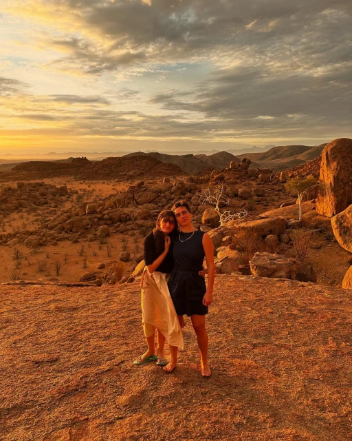 Two people pose on a rocky outcrop overlooking a desert landscape at sunset under a sky filled with orange and grey clouds.