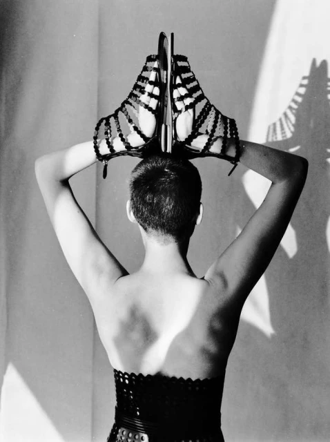 Back view of a person holding strappy gladiator sandals above their head against a white wall casting shadows. Fashion editorial.