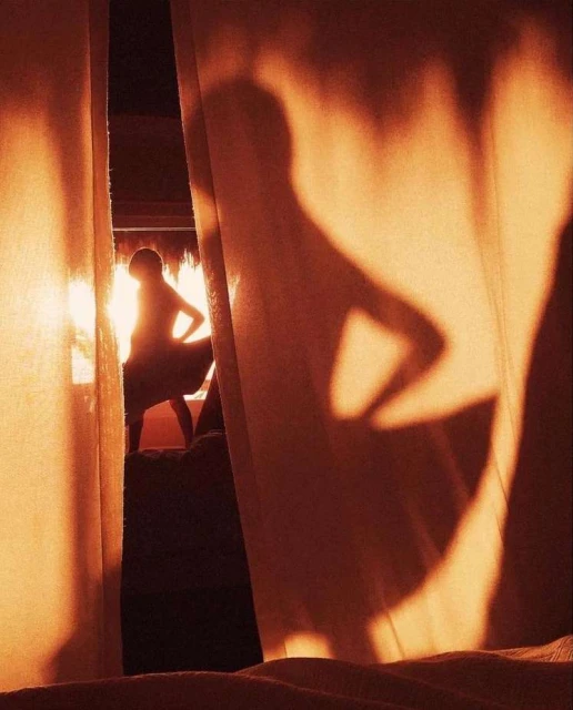 Woman's silhouette against bright window light, partially obscured by a curtain. Warm orange tones and a minimalist composition.