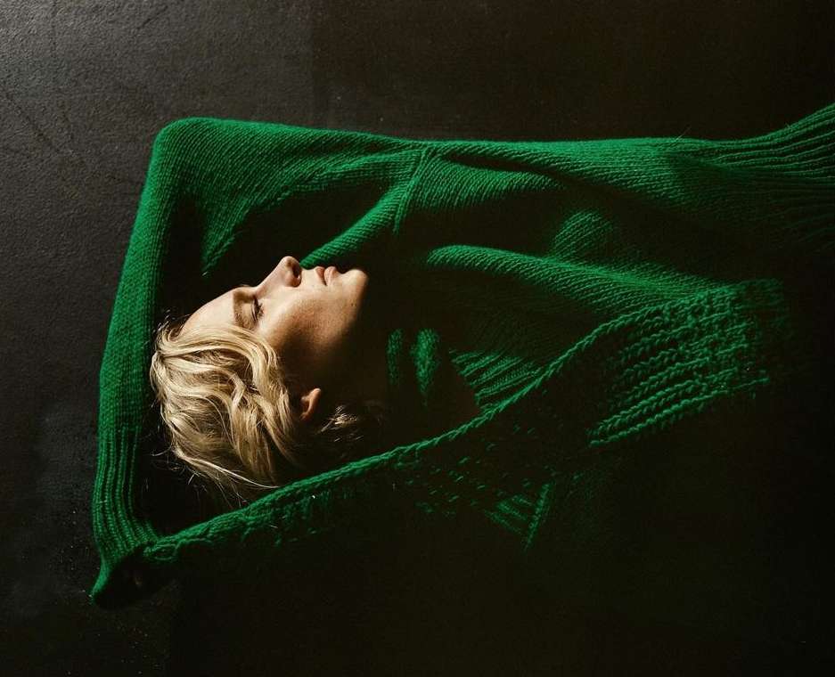 A woman lies on her back on a dark surface, wearing a vibrant green sweater, creating a stark contrast and a serene, cozy atmosphere.