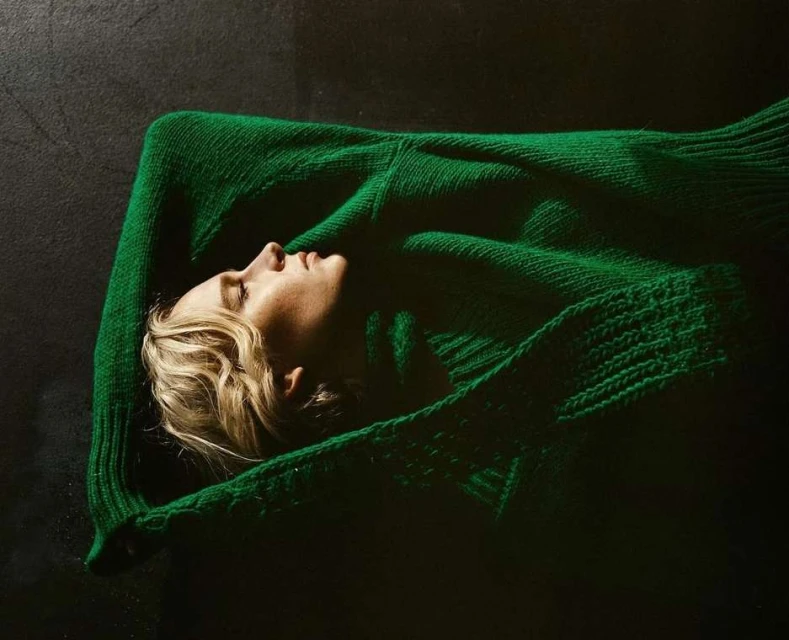 A woman lies on her back on a dark surface, wearing a vibrant green sweater, creating a stark contrast and a serene, cozy atmosphere.
