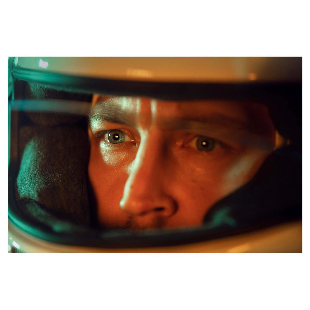 Close-up shot of a man wearing a racing helmet, lit with orange and green tones, creating a focused and intense portrait.