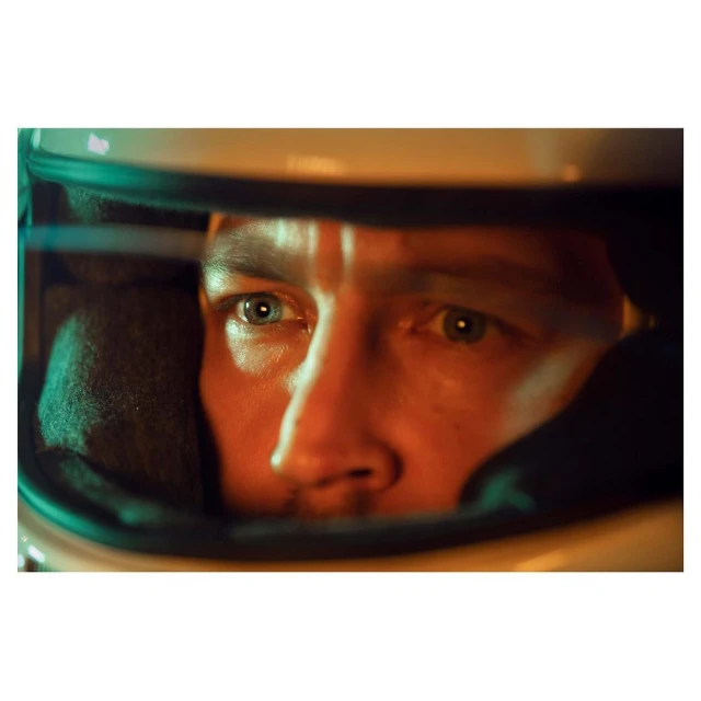 Close-up shot of a man wearing a racing helmet, lit with orange and green tones, creating a focused and intense portrait.