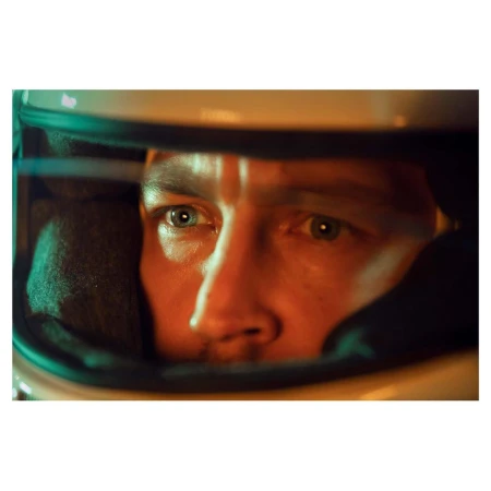 Close-up shot of a man wearing a racing helmet, lit with orange and green tones, creating a focused and intense portrait.