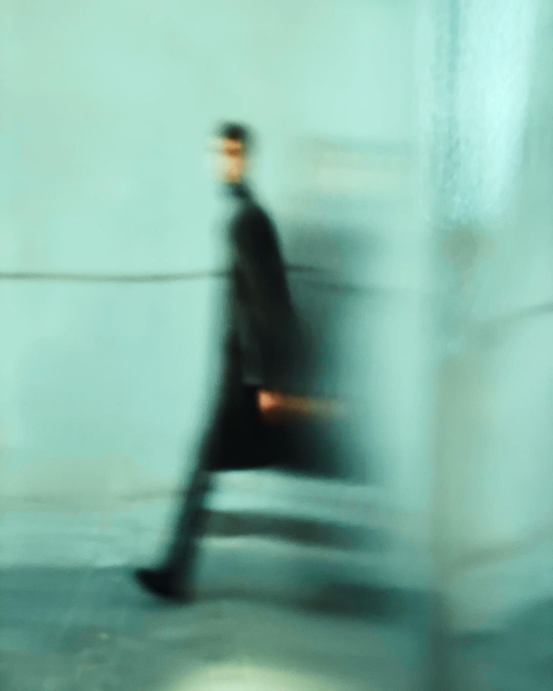 A blurry image captures a male figure in a long coat walking along a wall, conveying a sense of movement and modern urban style.