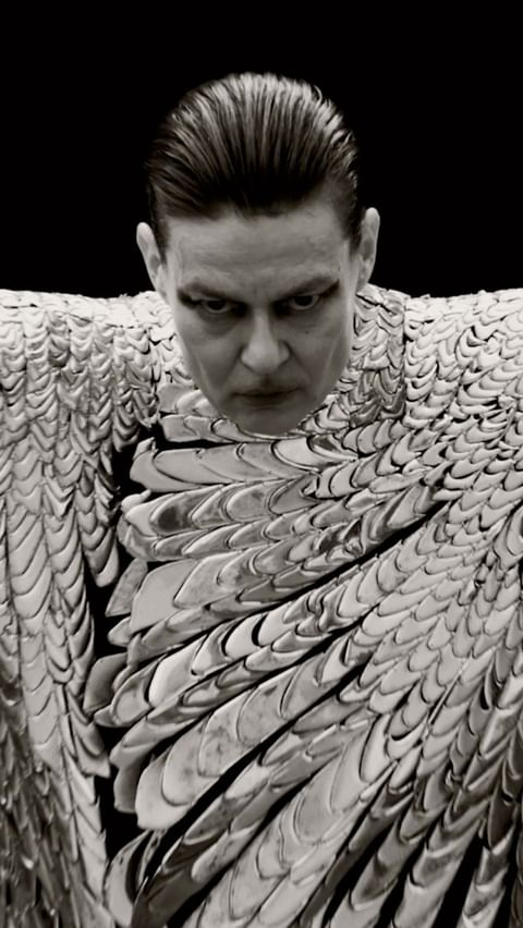 Monochromatic portrait of a man with slicked-back hair wearing a metallic scale-like garment against a stark black backdrop.