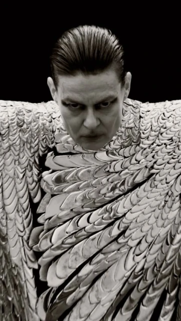 Monochromatic portrait of a man with slicked-back hair wearing a metallic scale-like garment against a stark black backdrop.