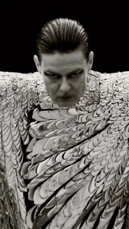 Monochromatic portrait of a man with slicked-back hair wearing a metallic scale-like garment against a stark black backdrop.