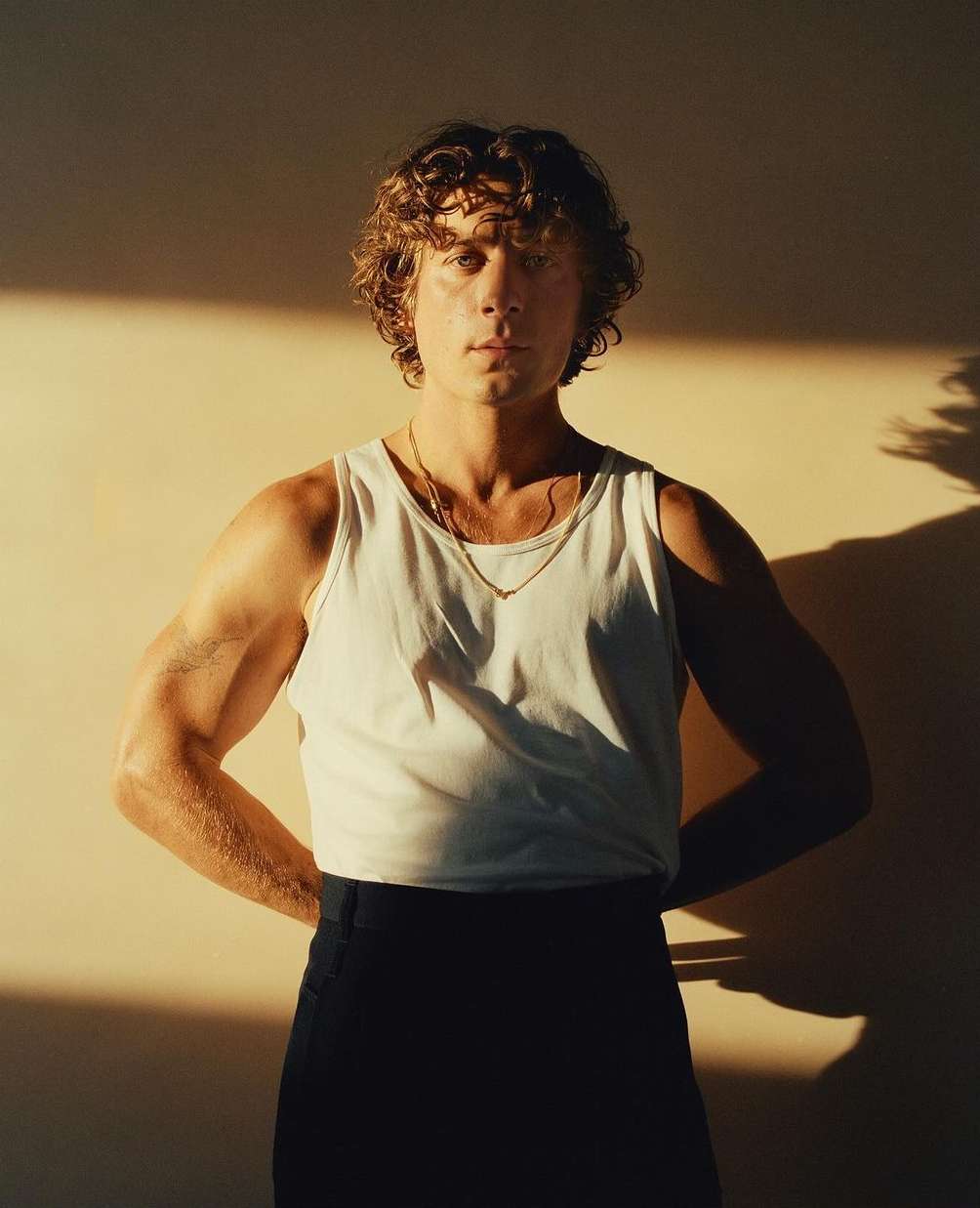Portrait of actor Jeremy Allen White with curly brown hair wearing a white tank top and dark trousers against a warm wall.