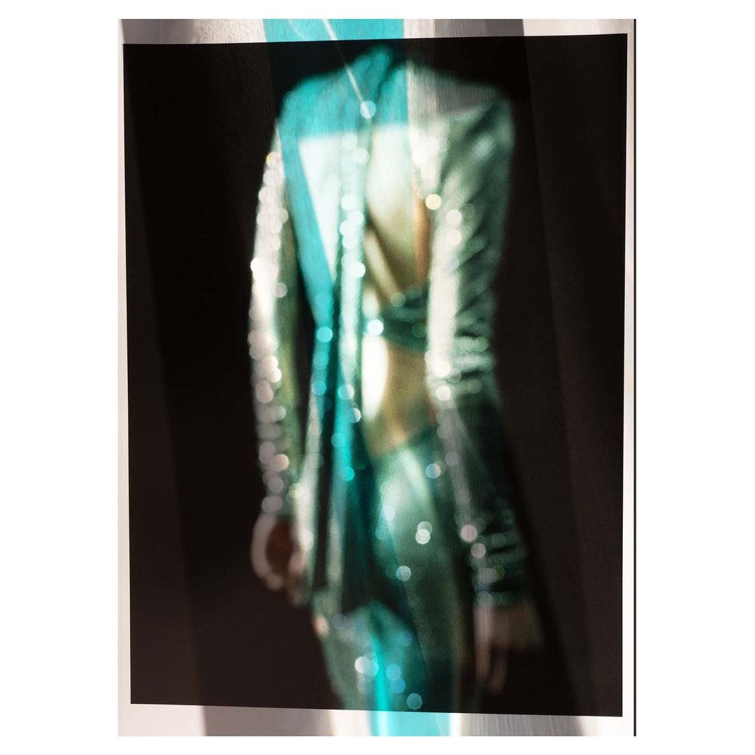 Woman in shiny teal outfit, visible from the rear in a blurry studio shot with dark background and diffused, even lighting.