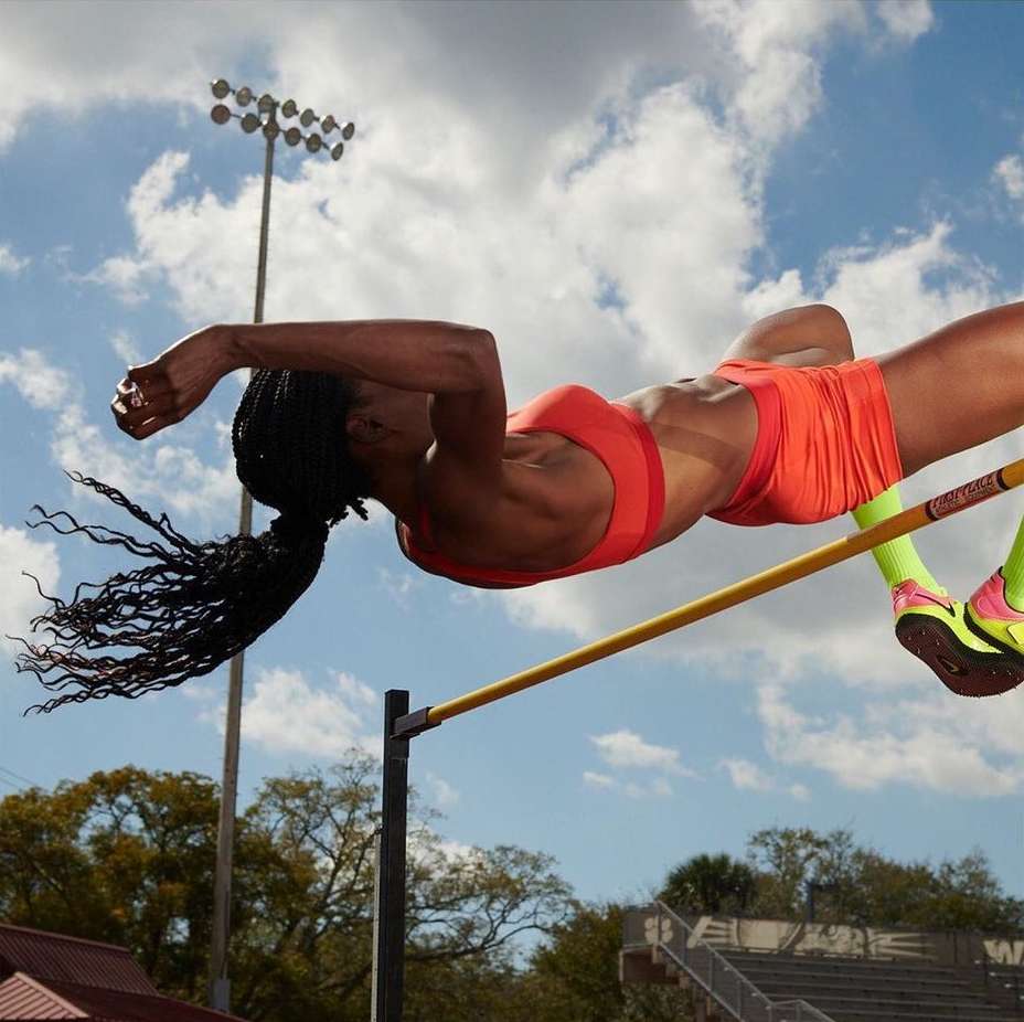 Female athlete in orange sportswear clearing the high jump bar outdoors under a blue sky with some scattered white clouds during daylight.