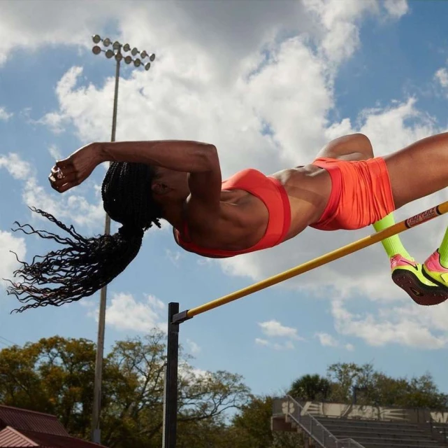 Female athlete in orange sportswear clearing the high jump bar outdoors under a blue sky with some scattered white clouds during daylight.