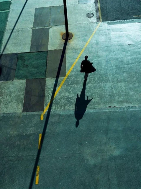 Aerial view of a solitary person walking on a concrete surface with a long shadow, next to a yellow line and a pole shadow.