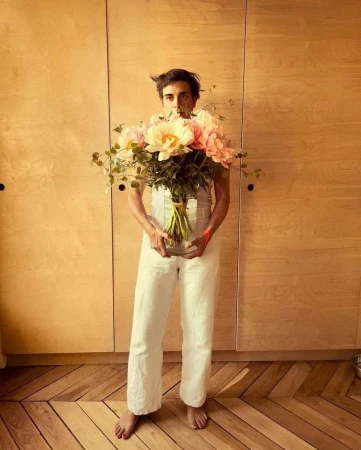A man standing barefoot wearing white overalls holding a large glass vase filled with large pink flowers in front of a wooden panel wall.