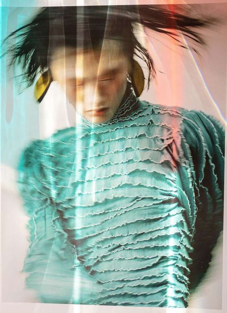 Fashion photo of an Asian adult female in a teal ruffled top and gold earrings, with soft lighting and a blurred, artistic effect.