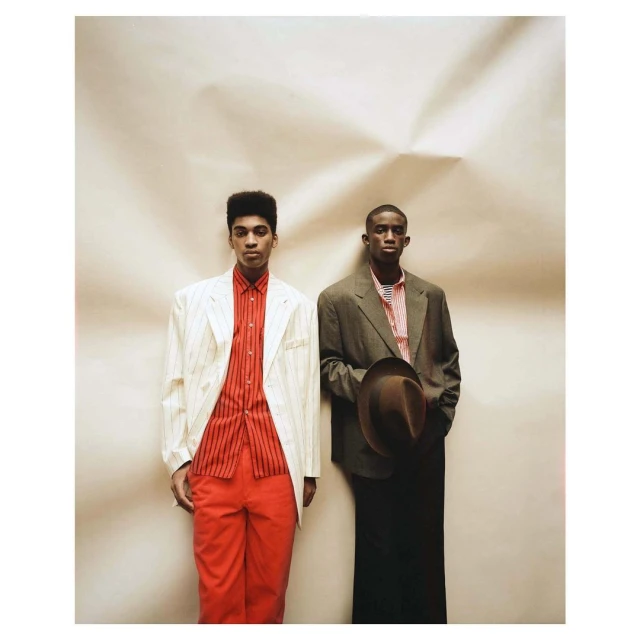 Two young adult men in suits, one in white, one in brown, stand in front of a mottled beige background, posing for a fashion shot.