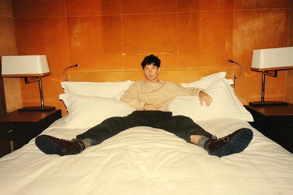 A man lying on a bed with a tan shirt, dark pants and brown boots, looking at the camera, with golden walls and lamps.