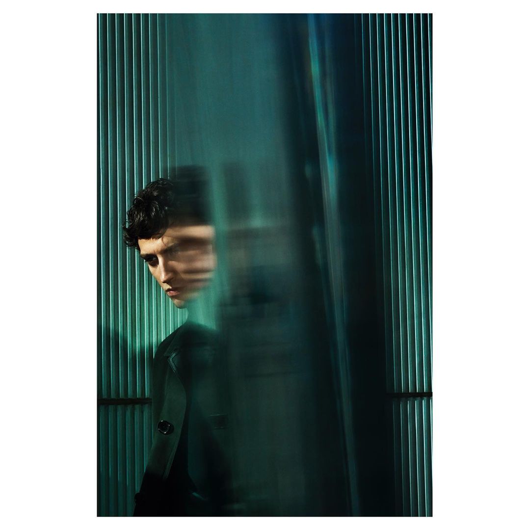 Portrait of a stylish young adult male model with dark curly hair, captured in a photo studio with teal lighting and an abstract background.