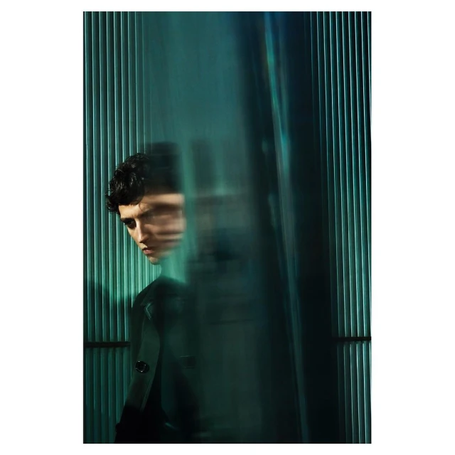Portrait of a stylish young adult male model with dark curly hair, captured in a photo studio with teal lighting and an abstract background.