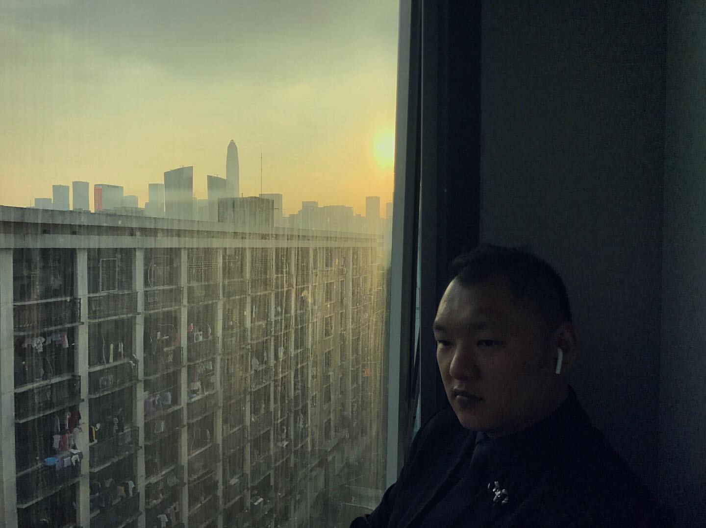 Adult male wearing a suit by a window overlooking an apartment complex and city buildings during a sunset, creating a serene and contemplative scene.