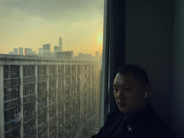 Adult male wearing a suit by a window overlooking an apartment complex and city buildings during a sunset, creating a serene and contemplative scene.