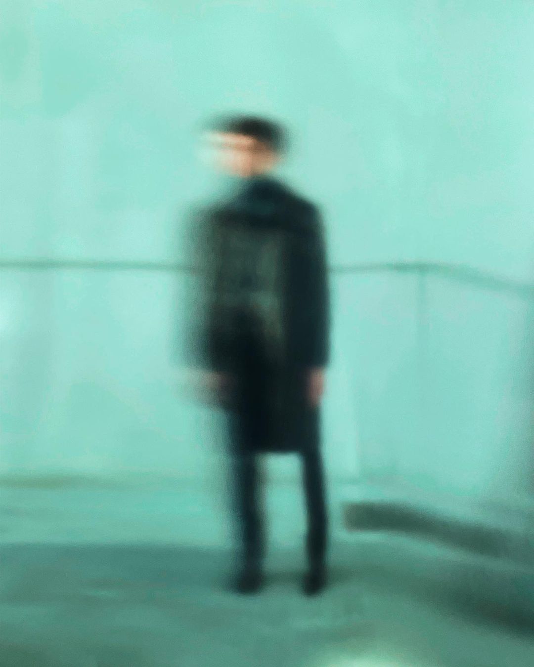 Male fashion model stands against an aqua wall, wearing a black overcoat and dark pants, captured with blur effect and low angle shot.