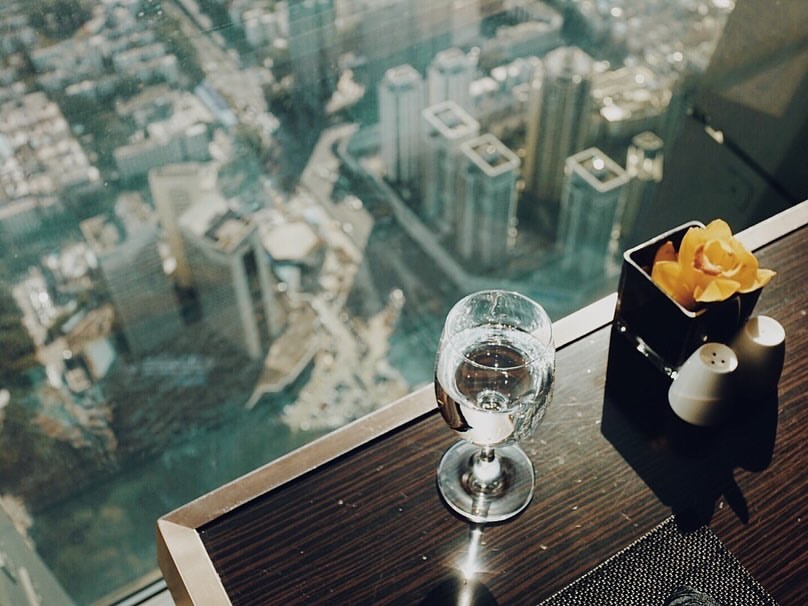 Aerial view of city buildings through window next to a glass of water and salt and pepper shakers on a wood table.
