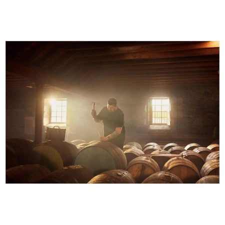 Adult man in rustic cellar hammering bung into wooden barrel in cellar with barrel collection, sunlight, woodworking tool in use.